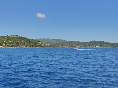 The image depicts a vibrant coastal scene under a clear blue sky with a single, fluffy white cloud. The foreground is dominated by the deep blue, rippling waters of the sea. In the mid-ground, a coastline rises, densely covered in lush green trees and dotted with white buildings, suggesting a populated area. A small, white motorboat with people aboard is speeding across the water, leaving a white wake behind it. The background features rolling, verdant hills stretching towards the horizon. While the specific location of Platanias, Greece, is not explicitly visible, the scenery is consistent with a Mediterranean coastal town.