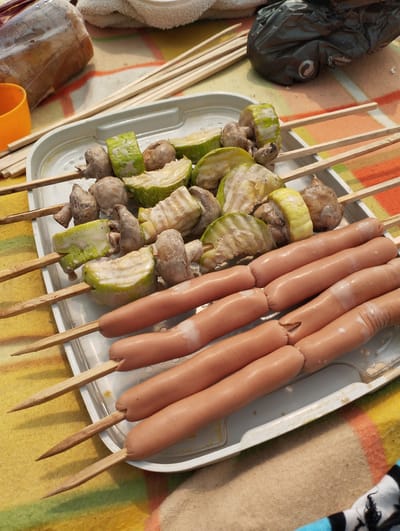 The image shows a white plastic tray filled with skewers of food, placed on a colorful striped blanket. The skewers are of two types: one type holds pieces of zucchini and mushrooms, some of which are coated in a white sauce. The other type holds sausages. The skewers are made of thin wooden sticks. In the background, there are more wooden skewers, an orange plastic cup, and a plastic-wrapped object that appears to be food. A black plastic bag is also visible in the background. The setting appears to be outdoors, possibly a picnic or barbecue, given the food and the blanket. The lighting suggests it is daytime. The overall mood is casual and suggests preparation for a meal. There is no visible text in the image.