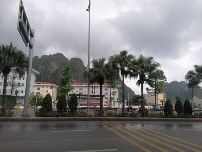 This image presents an overcast street scene in Ha Tu Ward, Vietnam, characterized by a wide, wet asphalt road in the foreground, suggesting recent rain. Yellow diagonal markings are visible on the road surface. A central median strip features neatly trimmed bushes, small evergreen trees, and several tall palm trees.
Beyond the median, a row of multi-story buildings lines the street, including a light blue structure on the left, a light yellow building further back, and a prominent greyish-white building with a red tiled roof. A large, metallic street sign or billboard frame is visible on the left side, and a very tall, slender pole extends into the cloudy sky from the center.
In the middle ground, two individuals on motorcycles are seen riding from left to right on the far side of the road, indicating everyday urban activity. Majestic karstic mountains, partially covered in greenery, dominate the background under a heavy, grey, cloudy sky.
A large billboard in front of the grey building displays Vietnamese text. Visible phrases include "XĂM HẠI PHỤ NỮ VÀ TRẺ EM LÀ TỘI ÁC NGHIÊM TRỌNG" (Sexual abuse of women and children is a serious crime) and "HÃY TỐ CÁO HÀNH VI XÂM HẠI BẠO LỰC ĐỐI VỚI PHỤ NỮ VÀ TRẺ EM" (Report acts of abuse, violence against women and children). The billboard also mentions "vinaphone" with a slogan that appears to be "SIÊU TỐC ĐỘ SIÊU TRẢI NGHIỆM" (Super speed, super experience). The daytime scene has a muted quality due to the heavy cloud cover.