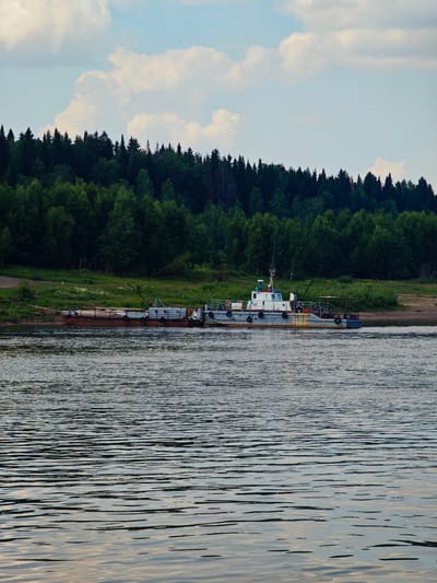 An external, daytime, vertical shot captures a broad, calm river with a barge and a small tugboat docked near the grassy bank. A dense, dark green forest lines the opposite bank, with a clear blue sky and scattered white clouds above. The barge, a long, flat structure with a rusted hull, is laden with what appear to be cylindrical objects. A smaller, white tugboat is moored alongside the barge, featuring a cabin, a mast, and railings. The river's surface is rippled, reflecting the subdued light. The overall scene depicts a quiet, natural environment with industrial elements.