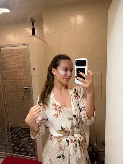 A woman stands in a bathroom and takes a selfie. She wears a floral robe over a top, and an eye patch on her left eye. She is holding a white smartphone. The bathroom has beige tiles, a shower with a sliding door, and a red curtain visible. A dark, cylindrical object with sticks is mounted on the wall. There is no evidence to suggest that the scene is located in Tarasovka, Russia.