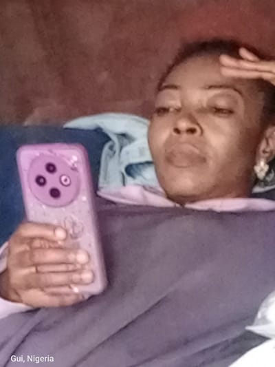 In Kuje, Nigeria, a medium shot captures a woman with her hand raised to her forehead, looking down at a purple smartphone held in her other hand. She is lying on a couch, covered by a purple blanket. A light blue sheet is visible behind her head. The smartphone has a distinctive camera module with four circular lenses arranged in a square formation. The background is a textured brown surface. The watermark "Gui, Nigeria" is present in the bottom left corner.