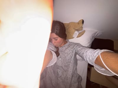 A person in pajamas lies in bed with a stuffed animal bear behind their head. One arm is extended towards the camera, showing an IV line on their arm. A bright light obscures part of the image. The scene appears to be taken indoors.