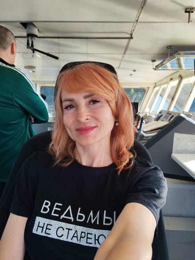 The image shows a medium shot of a woman with red hair and fair skin, wearing a black t-shirt with silver text that appears to be in Cyrillic script. She is smiling at the camera. She is in what appears to be the bridge of a boat or ship, indicated by the windows and control panels visible in the background. Another person, a man wearing a green tracksuit with white stripes, is visible in the background on the left, with his back to the camera. The environment suggests an indoor setting on a vessel, with natural light coming through the windows. There is no indication of the location being specifically in Pesochne, Russia.