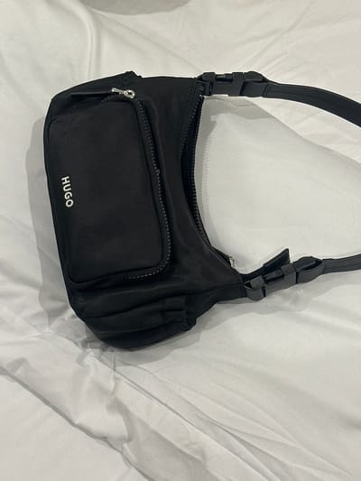 A black synthetic textile shoulder bag, featuring a white vertical "HUGO" logo on its zippered front pocket, lies diagonally on a white, rumpled fabric surface. The bag exhibits a main top zipper and an adjustable black strap secured by black plastic quick-release buckles. A silver-colored zipper pull is visible on the front pocket. The scene is located in Gillingham, United Kingdom.