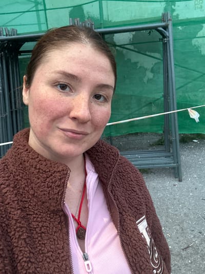 A close-up, slightly angled selfie features a young adult with medium-brown hair pulled back from their face, revealing freckles and reddish skin on their cheeks and forehead. They are wearing a brown, fuzzy jacket over a pink, ribbed top. A red string with a brown pendant hangs around their neck. A nose ring is visible. The background consists of a green tarp stretched over metal scaffolding. The ground is composed of gravel and dirt. The overall lighting suggests an outdoor setting during the day, possibly in an urban or construction area. There is no discernible location in Vilyuchinsk, Russia indicated within the image.
