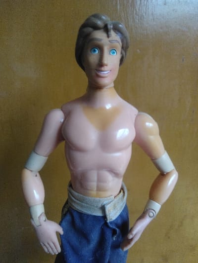 The image displays a plastic male action figure positioned vertically against a plain, textured, light brown background. The figure has sculpted brown hair, blue eyes, and a smiling mouth with visible white teeth. Its bare torso is muscular, molded in a light peach skin tone, and shows abdominal definition. Articulated arms feature light beige bands at the upper and lower arm joints. The figure wears dark blue fabric trousers with a light beige fabric belt around the waist. Its hands are open and relaxed, held slightly away from the body. The environment consists solely of the background, offering no visual cues to determine the location within the city of Saint-Gilles - Sint-Gillis, Belgium.