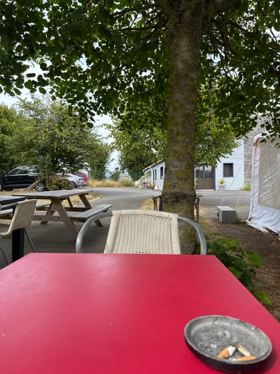 The image depicts an outdoor seating area with a red table in the foreground, an ashtray with cigarette butts, and a chair with a woven back. In the background, a picnic table and bench are visible, along with several parked cars. A tree with lush green foliage provides shade over the area. Beyond the trees, a white building with windows and a large white tent are situated. The ground is paved, and there is a patch of dry grass and some green bushes. The scene appears to be in Saint-Jouin-Bruneval, France, suggesting a casual outdoor dining or resting spot.