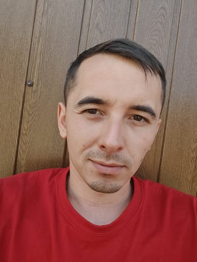 A medium-close shot shows a light-skinned man with short, dark hair, dark eyebrows, and light brown eyes against a wooden plank background. He is wearing a red t-shirt. A small, dark goatee is visible on his chin, and a thin chain necklace rests on his collarbone. The background consists of vertical wooden planks with visible grain and a single screw head. The image appears to be a selfie taken outdoors, possibly in a residential area of Shkurupiivka, Ukraine, given the context.