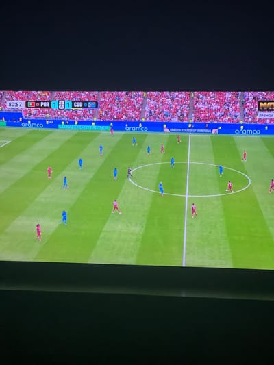 This image shows a screen displaying a soccer match. The score is 1-1 between POR and COD in the 80:57 minute. Several players in red and blue uniforms are on the green field. In the background, a stadium filled with spectators wearing red clothing is visible. Advertising banners for "aramco," "FIFA," and "GLOBAL ENERGY PARTNER" are displayed along the side of the field. The screen also shows the text "UNITED STATES OF AMERICA" and "МАТЧ ПРЯМОЙ". The overall scene is a broadcast of a live sporting event. The location of Bely Klyuch, Russia, is not discernible from the provided image.