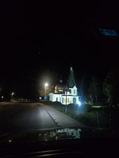At night in Svetlen, Bulgaria, a brightly lit white church with a green roof stands adjacent to a road. A triangular yield sign is positioned near the church. The road reflects the church's light and is lined with street lamps. Pedestrian crossing signs are visible along the roadside. A dark, unlit area extends to the left of the road and church. The image appears to be taken from inside a vehicle, evidenced by a partial reflection.