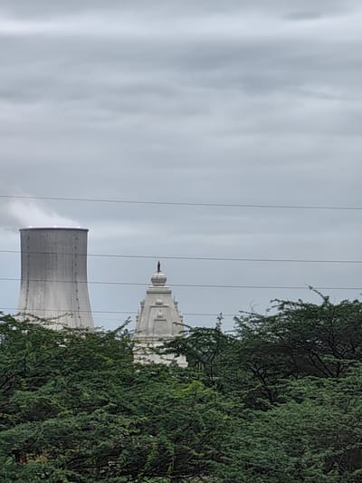 In Sudgaon, India, a wide shot captures a cooling tower of a thermal power plant on the left, emitting white steam from its top. To the right of the cooling tower, partially obscured by dense green trees, is a white temple structure with a distinctive tiered roof topped by a finial. The sky is overcast with gray clouds, and power lines are visible horizontally across the frame. The foreground is filled with the foliage of numerous trees.
