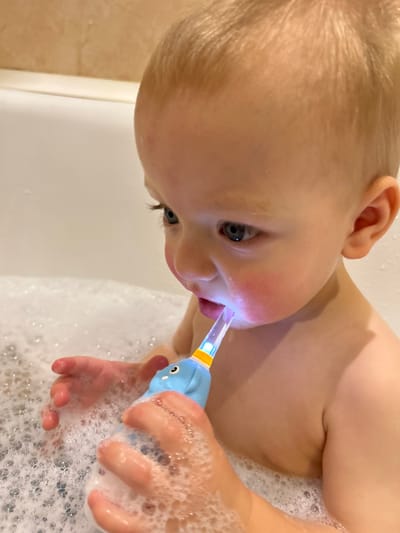 A young child with fair skin and blonde hair is in a bubble bath, holding a blue and yellow light-up toothbrush. The toothbrush is in the child's mouth, illuminating their face. The child's hand, covered in bubbles, grips the toothbrush handle. The background is filled with white, foamy bubbles and glimpses of the white bathtub edge. The image is taken from a slightly downward angle, focusing on the child's face and the illuminated toothbrush.