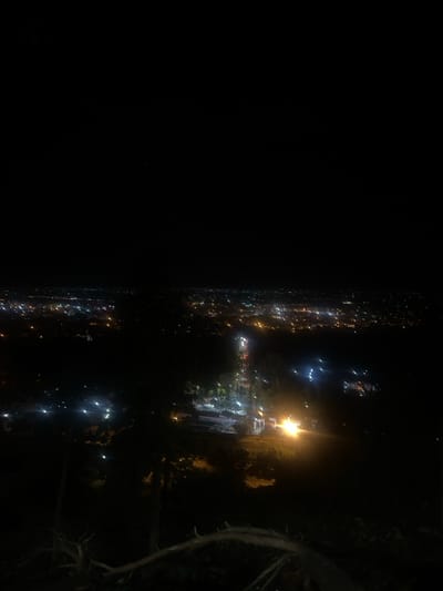 The image displays a panoramic nighttime view of Beni Mellal, Morocco, captured from an elevated perspective overlooking the city. In the immediate foreground, a dark, gnarled tree branch is visible, extending horizontally across the lower right portion of the frame, with other indistinct dark vegetation present. The midground features a section of the city with visible artificial illumination, including a winding road exhibiting streaks of red (taillights) and white/blue (headlights) from vehicles. Several brightly lit structures, consistent with buildings and public areas, are identifiable. The background is dominated by the sprawling urban landscape of Beni Mellal, characterized by a dense, extensive pattern of countless artificial lights, primarily yellow, orange, and white, extending towards the distant horizon. The sky above is uniformly dark. No individuals, specific actions, or interactions are discernible.
