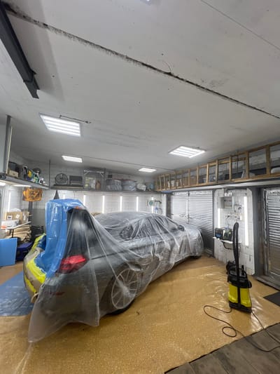 In a cluttered garage in Veteran-2, Russia, a car is covered in protective plastic sheeting, indicating it's undergoing some form of automotive service or detailing. The car, a dark sedan, is partially visible, with its rear taillight illuminated. The garage is brightly lit by fluorescent ceiling panels and strip lighting along the walls. Shelving units line the walls, filled with various automotive supplies, tools, and equipment, including a fan, cleaning bottles, and boxes. A yellow and black wet/dry vacuum cleaner is positioned on the floor near the car, connected to a power outlet by a black cord. The floor of the garage is covered with a light-colored, textured material, possibly a protective mat or coating, with some areas appearing damp. The overall scene suggests a professional automotive workshop actively engaged in work on the vehicle.