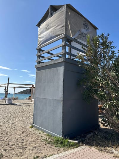 The image displays a sunny beach scene in Anatoliki Attiki, Greece, centered around a beach establishment. In the foreground, a prominent dark grey, box-like structure stands on the sand. Attached to this building is an open wooden framework that supports light-colored fabric awnings. These awnings are emblazoned with a distinct circular, geometric logo and the text "DIOPIO BEACH". A tree with green leaves, possibly an olive tree, grows alongside and slightly over the structure, casting some shadows.

The structure is situated on a sandy beach that extends towards the calm, clear turquoise sea. The sky above is a vibrant, cloudless blue, indicating bright and clear weather conditions, likely during the morning or early afternoon. In the distance, across the water, a low, undulating landmass or hill is visible on the horizon. Sparse beach furniture, including umbrellas and lounge chairs, can be seen on the sand further down the beach, suggesting a resort or beach club setting.

No people are visible in the image, contributing to a tranquil and serene atmosphere. The overall impression is one of a peaceful, sun-drenched coastal environment.