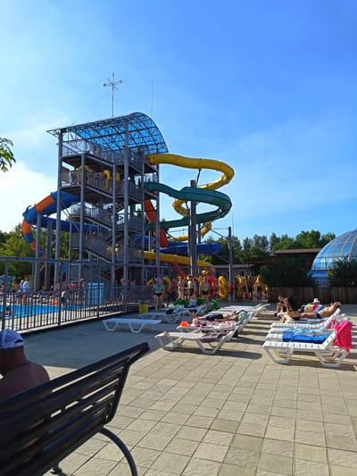 This media file depicts a sunny day at a water park in Yarovoye, Russia. The main subject is a large, multi-story water slide complex with blue, orange, yellow, and green slides. People are visible on the platforms of the slides, waiting to go down, and gathered at the base.
In the foreground, several lounge chairs are set up on a tiled patio area. Some chairs are occupied by people sunbathing, while others are empty. A dark metal bench with a person sitting on it faces away from the camera, towards the water slides. In the background, a swimming pool is visible with people in and around it. Beyond the water park attractions, lush green trees form a backdrop, suggesting a warm, pleasant day. A large, domed structure, possibly another attraction or building, is visible in the far right background.
The sky is a clear, bright blue, indicating fair weather and a time of day likely in the late morning or early afternoon, given the strong sunlight and shadows. The overall mood of the scene is relaxed and recreational, with people enjoying a day of summer fun. No visible text is present in the image.