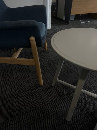 The scene is an indoor environment located in Dorchester, United Kingdom. The foreground features a dark grey carpeted floor composed of textured tiles arranged in an alternating linear pattern. On the left, a partial view of a dark blue upholstered armchair with light wooden legs and a visible frame is present. To the right of the armchair, a light grey circular side table stands on matching grey legs. In the background, a white door frame or archway is visible, leading to an adjacent area containing indistinct dark furniture and a white cable on the floor. No individuals, specific actions, or interactions are depicted.