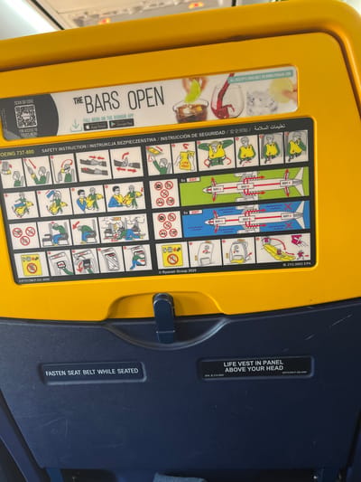 The image shows an airplane seat tray table in yellow and dark blue. The tray table has a safety instruction card attached, displaying safety instructions in multiple languages, including English, Polish, Spanish, Chinese, and Arabic. The safety card illustrates emergency procedures, like using a life vest. A sticker advertising "The Bars Open" and mentioning Ryanair is also present, with QR codes and links to download the Ryanair app for accessing the menu. Adjacent to the safety instructions are labels indicating "Fasten Seat Belt While Seated" and "Life Vest in Panel Above Your Head".

There is no indication of location (e.g., Casorate Sempione, Italy).