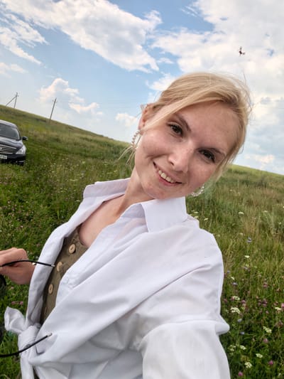 A blonde woman takes a selfie outdoors in a grassy field with scattered wildflowers. She is wearing a white open collared shirt over a green jumpsuit with wooden buttons. She has on large pearl earrings and is holding sunglasses. In the background, a black car is parked on the left side of the frame, and there are two utility poles with wires in the distance. The sky above is cloudy with patches of blue. There is a small insect flying in the upper right portion of the frame. The location is the outskirts of the village of Kuz'minovka, in the Belgorod Oblast, Russia.