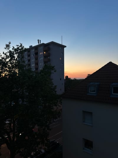 The image captures a twilight scene in Freiburg im Breisgau, Germany, with buildings and trees silhouetted against a gradient sky transitioning from a deep blue at the top to a warm orange and pink near the horizon.
In the foreground and right side of the image, a building with a dark, tiled, sloping roof dominates. This roof features two dormer windows with dark frames and what appear to be closed shutters on the left dormer. Below the roofline, the building's light-colored facade is visible, with two rectangular windows, one of which has closed shutters.
To the left, a taller, apartment-style building is partially visible. This building is gray and has multiple floors with numerous windows and balconies. Some of the windows on the balconies are lit, casting a warm glow. On the roof of this building, several antennas or communication towers are mounted. A slender antenna protrudes from the roof on the far left.
A large, dark silhouette of a tree with dense foliage is positioned in the middle left of the frame, obscuring a portion of the apartment building and the street below. The street, visible beneath the tree and in the background, appears to be an urban road with traffic markings. Several vehicles, including cars, are present on the street and parked along its edge, some partially obscured by the darkness and the tree.
The overall environment suggests a residential or urban area during the evening transition from day to night. The sky indicates a clear or partly cloudy condition with the sun having recently set.