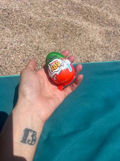 A hand holds a Kinder Joy Minecraft edition chocolate egg over a textured teal surface. The hand has a tattoo of the number "13" on the wrist, on the inner side of the left arm. The background consists of small, light-colored pebbles, indicative of a beach. The lighting suggests it is daytime. The location is the beach in Sant Pol de Mar, Spain.