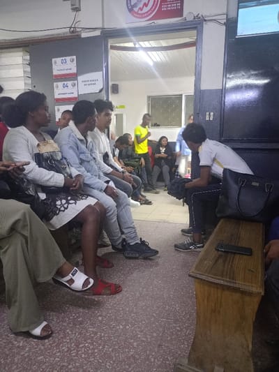 The image shows a waiting room in Antananarivo, Madagascar (latitude: -18.917, longitude: 47.517). The room is brightly lit and appears to be a government or healthcare facility. A group of people sit on chairs lined against a wall, while another person is seated on a wooden bench in the foreground. The room has tiled floors, white walls with red and black signs, and an open doorway to another room. A woman in a white dress and sandals sits on a chair, holding a black handbag. She is seated next to a man in a blue denim jacket. Both appear to be waiting. Across the room, a man in a white t-shirt and blue jeans sits on a chair. He is looking down. Next to him, another young man in a white shirt is looking towards the entrance. A black handbag is lying on a wooden bench. A black cellphone is lying on the wooden bench. A man wearing a white t-shirt with black lettering, black pants and black shoes stands in the doorway. A woman in a white dress and red sandals is seated on a chair next to the man in the blue denim jacket. A young man in a white uniform and dark pants sits on the wooden bench. The young man in the white uniform and dark pants is talking to a man seated next to him on the bench. The man next to the young man is wearing a dark shirt and dark pants. He is looking down and not smiling. A woman in a white dress and white sandals sits on a chair, holding a black handbag. She is seated next to a man in a blue denim jacket. They appear to be waiting. A woman in a yellow dress and black shoes stands in the doorway. The scene is a waiting area in a government or healthcare facility. The image does not provide any information about the activity or the reason for the gathering.