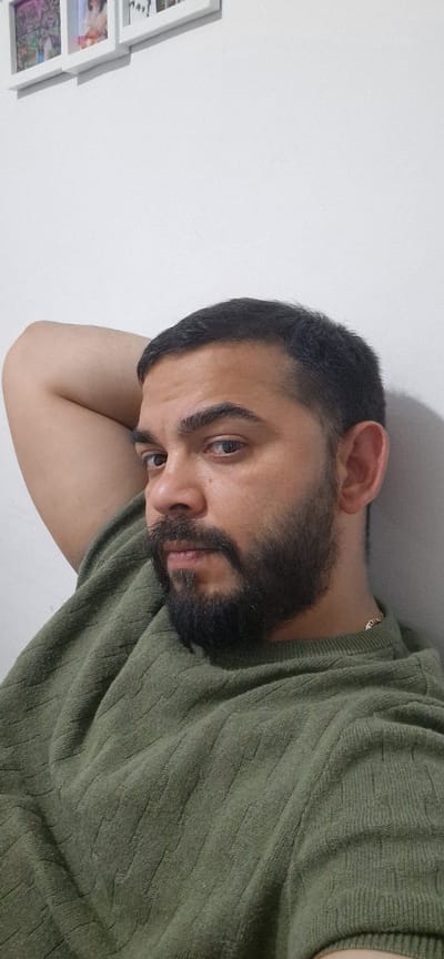 The image is a rotated close-up portrait of a man, likely a selfie, taken indoors. The man has short dark hair, a full dark beard, and a mustache. His eyes are light-colored, and he looks directly towards the viewer with a neutral, slightly pensive expression. He is wearing a dark olive-green sweater with a prominent ribbed or patterned texture. A thin gold chain is visible around his neck. His head rests on his bent arm, which is partially visible in the lower foreground. The background features a plain white wall. On the left side of the frame, a stack of white-framed pictures or photos is partially visible on the wall. The lighting is even, suggesting an indoor setting. There is no visible text in the image.