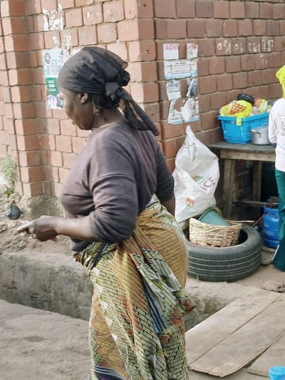 A woman in a black headscarf and long-sleeved gray shirt stands next to a brick wall and a concrete surface. She wears a colorful patterned wrap around her waist. Her right hand is visible, holding a cigarette. Behind her are various items including a tire, a blue basket, a white bag labeled "ICE," and other objects partially obscured. Torn posters are visible on the brick wall. The scene appears to be outdoors in a somewhat cluttered environment.