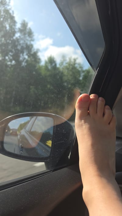 In an exterior shot of a car interior, a bare foot is visible, propped on the car door frame near the side mirror. The foot is light-skinned with visible toes and toenails. The side mirror reflects a blurry image of a car and trees. Outside the car, the sky is blue with scattered clouds, and trees are visible through the window. The interior of the car appears to be made of black textured plastic. The image is taken during the day, with sunlight creating shadows and highlights. The specific location in Kruglikovo, Russia is not discernible from the visual information provided.