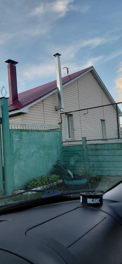 A low-angle shot captures the side of a beige, vinyl-sided house with a dark red metal roof. Two chimneys rise from the roof: one is a dark red rectangular structure on the left, and the other is a taller, silver metal pipe on the right. A green metal fence, partially obscured by a taller, solid green section, runs across the foreground. Behind the fence, a sparse garden with green plants is visible. To the right, a chain-link fence runs parallel to the solid green fence. The scene is set against a pale blue sky with scattered white clouds. In the bottom foreground, a dark gray dashboard is visible, with a small black cylindrical air freshener labeled "ORGANIC" placed on it. The location is identified as Tashkazgan, Russia.
