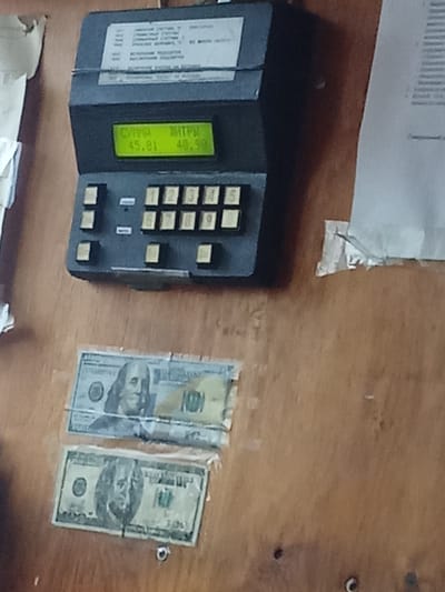 In Alatau, Kazakhstan, a wooden wall displays a black electronic device with a green LCD screen. The screen shows Cyrillic text and numbers: "СУММА 45.81" and "ЛИТРЫ 40.90". Below the screen are rows of buttons, some with numbers and others blank. To the left of the device is a partially torn piece of paper. Below the device, two U.S. one-hundred-dollar bills are taped to the wall. The top bill has a clear plastic protective sleeve, and the bottom bill is partially covered by another clear plastic piece. A small metal screw is visible on the wooden surface below the bottom bill. The lighting suggests an indoor environment with ambient light.