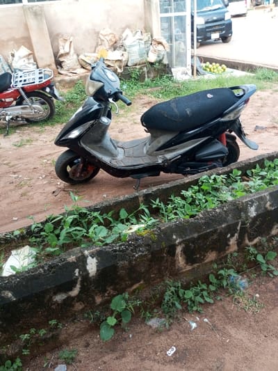 The image displays an outdoor scene in Oko Town, Nigeria. In the mid-ground, a black scooter with a black mesh seat cover is parked on its kickstand on unpaved reddish-brown dirt. A red motorcycle is partially visible behind the scooter on the left. In the foreground, a concrete drainage ditch, heavily overgrown with green vegetation and exhibiting dark stains, traverses the frame. Small pieces of white debris and a blue plastic bottle are visible within and around the ditch. In the background, a light-colored building wall is visible, with several large, light-colored sacks stacked against it. To the right of the sacks, a glass-paneled door or window is present. Further back on the right, a dark-colored minivan or small truck is partially visible on a paved surface, with a pile of green spherical fruits arranged on the ground near its front. No individuals are present.