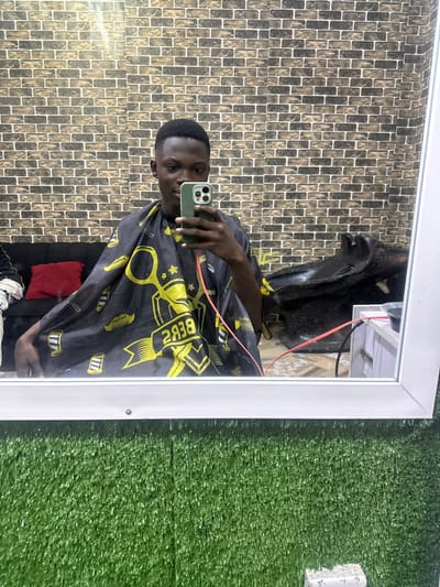 A man in a barber's cape, reflected in a mirror, holds up a phone taking a selfie. The cape is black with yellow graphics. The background features faux brick wallpaper on one side and artificial turf on the other. The mirror has a white frame. A barber's chair is visible in the reflection.