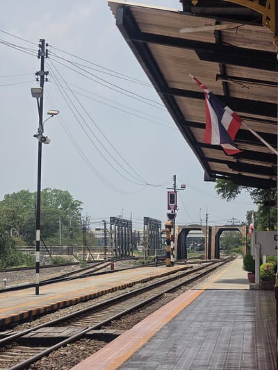 The image shows a view along a railway platform in Phra Nakhon Si Ayutthaya City Municipality, Thailand. The platform is lined with yellow and black striped safety markings. Train tracks run parallel to the platform. Overhead, there is a corrugated metal roof structure. The Thai flag is prominently displayed. A utility pole with wires and a loudspeaker stands adjacent to the platform. In the background, there are signal towers and other railway infrastructure. Beyond the infrastructure, trees and foliage are visible. The sky is overcast.