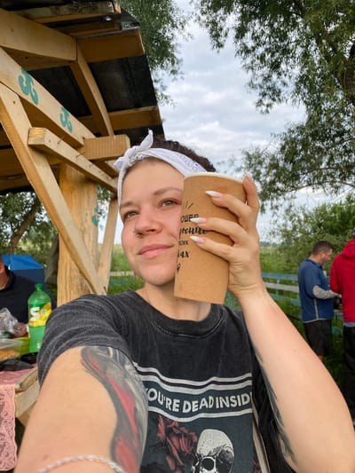 In Petrovskoye, Russia, a young woman with a white bandana tied around her head and a nose ring takes a selfie outdoors. She is holding a disposable coffee cup in front of her face, obscuring part of it. She is wearing a dark t-shirt with the words "YOU'RE DEAD INSIDE" printed on it, along with a graphic of a skull and roses. She has a large tattoo on her left arm, partially visible. In the background, a wooden structure with a tin roof is visible, as are trees and a cloudy sky. To the left, a green bottle of Sprite and some food are on a table. In the further background, two men are standing near a blue tent and a fence. The setting appears to be a park or a campsite.