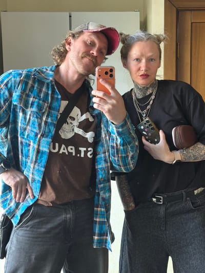 Two people are taking a selfie in front of a mirror. One person, presumed to be male, is wearing a baseball cap, a brown t-shirt with a skull and crossbones design, a blue plaid shirt, and dark jeans. The other person, presumed to be female, is wearing a black shirt, grey jeans, and several chains around their neck. Both people have visible tattoos. The male is holding an orange phone, and the female is holding a black phone with stickers on the back. The background appears to be an interior space with light-colored walls. There is no indication that the location is in Тэльмінскі сельскі Савет, Belarus.