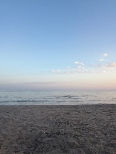 The media file depicts a serene beach scene at Cambrils, Spain. The composition is dominated by a vast expanse of clear, light blue sky occupying the left two-thirds of the frame. A scattering of small, wispy clouds, tinged with soft pink and white, are visible along the horizon where the sky meets the sea. The sea itself forms a narrow strip of muted blue water, with subtle ripples indicating gentle movement. To the right, a textured sandy beach fills the remaining portion of the image. The sand appears damp and somewhat coarse, with visible imprints and variations in tone, suggesting it has recently been exposed to the tide or wind.

The image captures a tranquil moment, likely during sunrise or sunset, given the soft, diffused light and the colors in the sky. There are no people or man-made objects immediately visible, contributing to the peaceful, natural atmosphere. The orientation of the image is landscape, but it has been captured in portrait orientation, so the scene is sideways. The overall impression is one of calm and natural beauty.