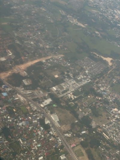 This aerial photograph offers a high-angle view of a mixed urban and rural landscape in San Phra Net, Thailand. The scene is characterized by a sprawling arrangement of buildings, roads, and tracts of land.
The subjects primarily include a dense network of residential and commercial structures, identifiable by their varied roof colors and rectangular forms. These buildings are often arranged in grids, forming discernible neighborhoods and developments. Interspersed among these built-up areas are patches of green vegetation, indicative of trees or agricultural fields, and lighter, earthy-toned areas which could be undeveloped land, dry ground, or construction sites.
A significant feature is a multi-lane highway cutting across the middle-right and bottom-right sections of the image, with what appear to be vehicles traveling along it. Numerous smaller roads and dirt paths connect different parts of the visible area. A distinctive, irregularly shaped light-colored area, possibly a dry riverbed or a large cleared tract of land, is visible in the upper-middle left.
The image is captured during daylight hours, though the exact time is not discernible due to the soft, diffused light and the presence of atmospheric haze, which lends a muted quality to the colors and somewhat blurs fine details. There are no discernible people or readable text visible within the frame. The overall impression is one of an expanding settlement situated amidst natural or agricultural surroundings.