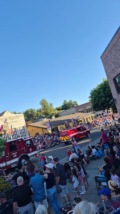 The image depicts a vibrant street parade, likely a Fourth of July celebration, with a large crowd gathered on both sides of the street. People are seated in chairs and standing, many holding American flags. The parade features a bright red fire truck adorned with flags, which is the central object in the image. A second fire truck is partially visible at the bottom of the frame. The scene is set on a sunny day, indicated by the clear blue sky and the shadows cast on the ground. Buildings line the street, with one featuring a sign that reads "WELLS FARGO & CO. BANK OFFICE". The atmosphere appears festive and patriotic, with many onlookers dressed in red, white, and blue. The time of day seems to be late afternoon or early evening, given the quality of light. The overall mood is one of community engagement and celebration.