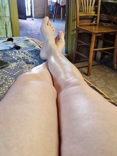 In Raduvene, Bulgaria, a low-angle shot focuses on the legs of a person resting on a patterned blanket or rug. The person's legs are bare and appear to be slightly oiled or moisturized, reflecting light. The feet are visible, with the toes of the left foot slightly curled. The background suggests an indoor setting with a rustic ambiance. A doorway leading outside reveals a textured wall and a patterned rug on the floor. To the right, a wooden chair with a spindle back is positioned in front of a wall adorned with a Greek key pattern. The lighting is natural, casting soft shadows.