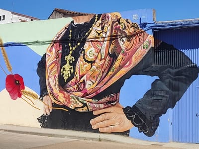 This is a mural painting on a building's exterior wall, located in Valencia de Alcántara, Spain. The artwork depicts a close-up of a woman's torso and arms, adorned in traditional Spanish attire. She wears a black dress with intricate black lace trim on the sleeves and collar. A vibrantly colored shawl, featuring intricate paisley and floral patterns in shades of pink, orange, yellow, and brown, is draped around her shoulders and chest. A prominent gold necklace with dangling charms hangs over the shawl.
In her left hand, held near her waist, she holds a single red poppy and two stalks of wheat, symbolizing nature and perhaps harvest or remembrance. Her right arm extends downwards, with her hand resting on her hip. The background is a split of colors: a light green band across the top, transitioning to a bright blue behind the woman's figure. To the right, a blue corrugated metal door and wall are visible, with a light blue electrical box mounted on the wall.
The mural is illuminated by bright daylight, casting shadows on the corrugated door. The sky above is a clear, vibrant blue. The mural has text painted on the blue wall near the top right corner, which appears to read "BREA", "ACUA", and "DROS" within a square. The overall impression is one of cultural heritage and artistic expression, set against a sun-drenched Spanish backdrop.