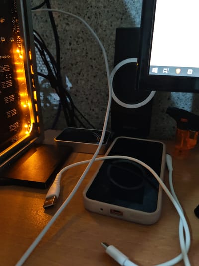 The image shows a desk with a computer tower, a monitor, a smartphone, and a charging cable. The computer tower has orange lights and is positioned on the left side of the image. A monitor is on the right side, displaying a white screen with some icons at the bottom. A smartphone with a light-colored case is in the center, with a white charging cable connected to it. The cable extends to a USB port, which is also visible. In the background, there is a black speaker with a white ring and a small orange lighter. The overall environment appears to be a dimly lit room, likely an office or a study. The location in the city of Ucar, Azerbaijan is not evident from the image.