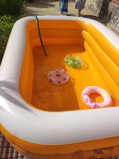 This media file shows an elevated view of an empty, inflatable, orange and white swimming pool set up outdoors, likely in a residential backyard in Mingchinar (Blagoveshchensky), Uzbekistan. The pool has a grid-like pattern on its orange floor. Three inflatable swim rings are visible inside the pool: one green with a cartoon animal pattern, one translucent pink with cartoon characters and bees, and one pink with a mermaid design and the text "The Pearl Princess". A green hose is draped over the edge of the pool, suggesting it's either being filled or has just been used to fill the pool.
In the background, two individuals are partially visible, wearing brightly colored dresses. Their legs and feet suggest they are standing on a paved surface next to the pool. The lighting indicates it is daytime, likely sunny, casting shadows from the pool and people onto the ground. The presence of lush green foliage to the left of the pool suggests a garden or green space nearby. The overall scene conveys a sense of leisure and preparation for outdoor activities, possibly related to a warm day. The text "TEPLO" is visible on the floor of the pool.