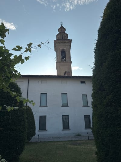 The image shows a view of the bell tower and a white building, possibly part of a monastery or church, in Balerna, Switzerland. The bell tower, made of stone, stands tall against a clear blue sky with a few scattered clouds. The white building features several rectangular windows, some with decorative grating. In the foreground, lush green trees and some leafy branches partially obscure the view, suggesting a garden or park-like setting. The overall scene conveys a sense of peacefulness and architectural grandeur.