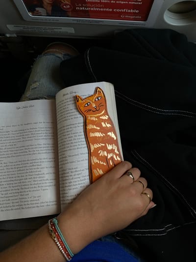 The image captures a top-down, angled view of a person reading a book, likely while seated on an airplane. The main subject is a person's right hand and arm holding an open book. The hand wears two gold rings, and the wrist is adorned with multiple bracelets, including beaded turquoise, red braided, and silver/rhinestone bands.
The open book features a distinctive orange cat-shaped bookmark with gold striped details, positioned at the top of the page. The text on the pages is legible in places, mentioning character names such as Mick, Nina, Kit, Ashley, and Jay, and phrases like "daughter's anger" and "elephant in the room," indicating a narrative story.
The person is wearing blue jeans with some distressed areas and a black jacket or top is draped nearby, with visible white stitching. To the left, an airline seat back or tray table is visible, along with an informational card. This card contains bilingual text in Spanish and English: "ABROCHE SU CINTURON DE SEGURIDAD MIENTRAS ESTE SENTADO" and "FASTEN YOUR SEAT BELT WHILE SEATED," confirming an aviation setting. Below this, another advertisement card is visible, promoting a "Laxante 100% de origen natural" (100% natural laxative) with the brand "Megalabs" and a website "www.planetite.com/viva". The overall setting is indoors, with no clear indication of the time of day.