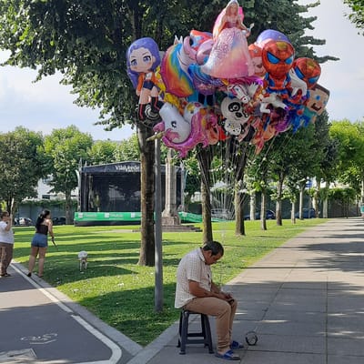 In Vila Verde, Portugal, a man sits on a stool by a paved path, looking at his phone. He wears a checkered shirt and tan pants. A kettlebell rests on the ground near his feet. To his left, a cluster of character balloons, including Spider-Man, a unicorn, a panda, and a princess, are tethered to a pole. Further left, two women and a small dog are on a grassy area. One woman is walking the dog on a leash, while the other stands nearby. In the background, a stage with the "Vila Verde Município" logo is set up in a park-like area with trees. Cars are parked along the road beyond the park.