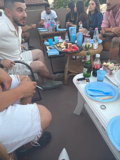 The image depicts a rooftop gathering at dusk, possibly in Troyanovo, Bulgaria, as suggested by the overall ambiance of an outdoor social event with a tiled roof visible in the background. The scene is centered around a white table laden with food and drinks, and several people are engaged in conversation and relaxation.
In the foreground, a person with short dark hair and a beard, wearing a white linen shirt and white ripped denim shorts, is seated with a cigarette in hand, appearing to be mid-conversation. To their right, another individual, partially visible, also in white attire, is seated.
Further back, a man in a white t-shirt is seated at a wooden table, holding a pink cup. Adjacent to him, a woman with dark hair and a black top is smoking a cigarette and looking towards the left. Another woman, wearing a dark blue top, is seated and appears to be looking down. On the far right, a person in a pink shirt with visible tattoos is seated.
The white table holds various items, including a platter of sliced watermelon, skewers of fruit and cheese, a bottle of Coca-Cola, a green bottle, several blue cups, a pitcher with lemon and cucumber, a salt and pepper shaker, and a pack of cigarettes with a lighter. A blue plate with a blue fork and wooden skewers is also present on the table. On a lower white table, there are more blue plates and a bottle cap. The flooring appears to be dark grey. The lighting suggests it is either early morning or late evening.