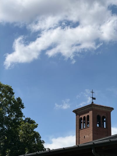 The image depicts a low-angle view of a brick clock tower against a bright blue sky with scattered white clouds. A weather vane with an arrow and the letters "SA" is visible on top of the tower. The clock tower has arched openings, suggesting it houses bells. To the right, a portion of a dark roofline juts into the frame, and below the sky, the lush green foliage of a tree is visible. The image appears to be taken during the daytime, with clear weather conditions. The style of the clock tower suggests a historical or religious building, and the presence of the weather vane might indicate a connection to navigation or a specific architectural style. The overall scene evokes a sense of historical architecture and a pleasant, clear day. The location is Piacenza, Italy.