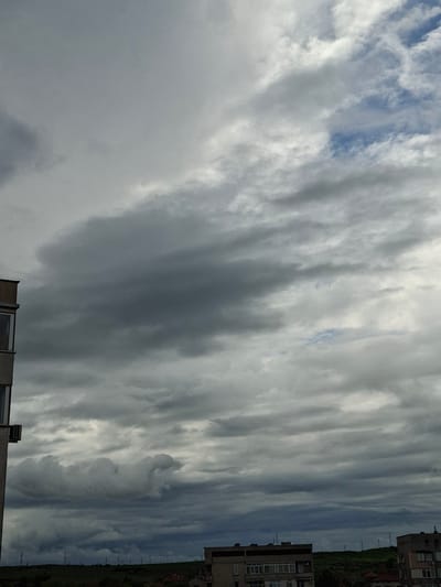 The photograph depicts a cloudy sky over Galabovo, Bulgaria. The sky is predominantly covered with gray and white striated clouds, with small patches of blue visible between them. A portion of a building is visible in the foreground at the bottom of the frame. On the right edge, buildings are present next to a green area with power lines. The orientation of the photograph is vertical.
